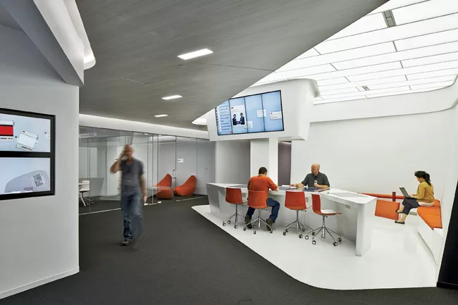 Five different “hubs,” each coded by a particular color, offer unprogrammed spaces for people to meet or relax. Overhead monitors and screens embedded in the work surfaces keep employees i
