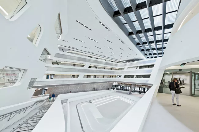 In Hadid’s building, a pair of canted volumes containing the library and the Learning Center shift around and define a central atrium.