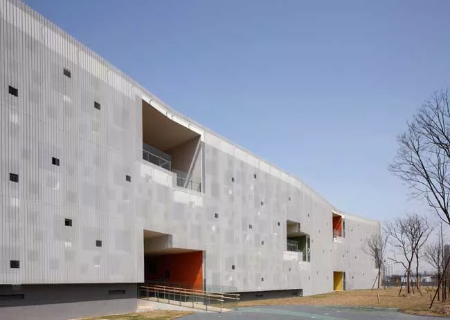 In the kindergarten, two parallel structures play in opposition to each other. A formidable concrete block encloses the circulation ramps for the school, while a punctured block with perforated alumin