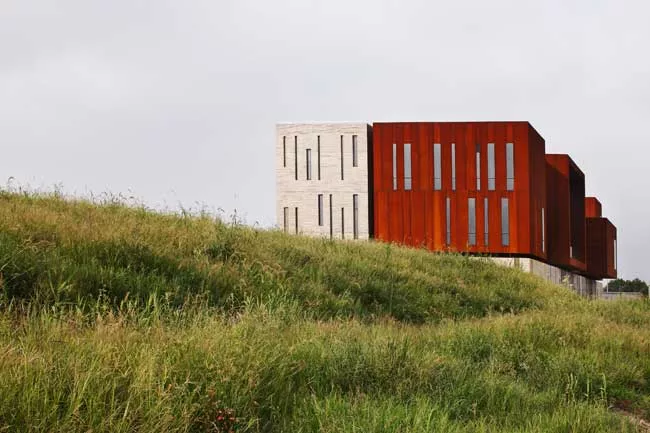 Surrounded by roads and vacant lots on three sides, the site for this building has just one notable feature: A river that runs along its north side. On its short sides, the building appears as a pair 