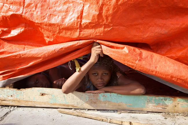 Young girls peek out from under a tarp shelter in a refugee camp in downtown Port-au-Prince. <em>April 2010</em>.