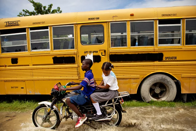Kids navigate floodwaters on a small motorcycle, passing a yellow school bus. <em>April 2010</em>.
