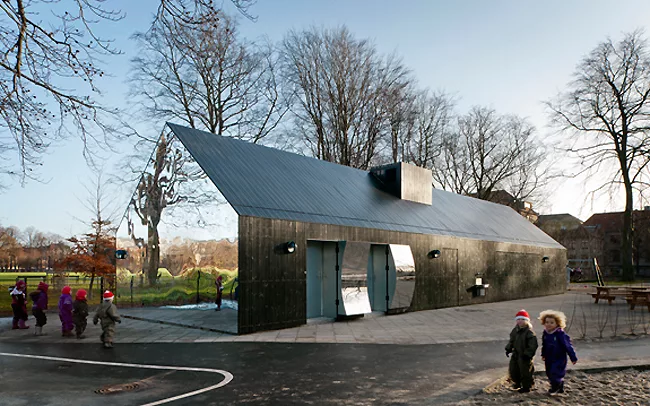 Positioned next to a playground, children approach the concave and convex mirrored surfaces on the gabled ends and behind the doors on the sides of the building to look at their distorted images.