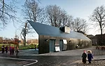 Positioned next to a playground, children approach the concave and convex mirrored surfaces on the gabled ends and behind the doors on the sides of the building to look at their distorted images.