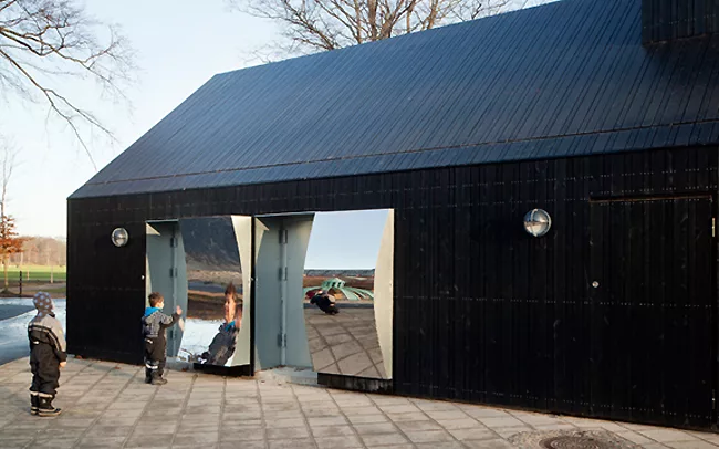 Thick doors open to reveal mirrors on their backsides, composed of polished stainless steel. The doors provide access to the inside of the building, where bathrooms, a playroom, a kitchen, areas for e