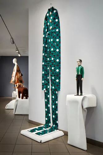 A stretched men’s suit by John Bartlett, based on a late 19th century wood statue, evokes both Joseph Beuys and a car dealership’s windsock. Yeohlee Teng’s paper dress is in the back