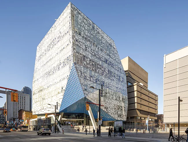 The building's distinctive form and the blue shard of the entrance canopy visually mark the gateway to Ryerson University amid the retail patchwork of the busy downtown street.