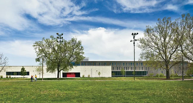 On the west elevation, the light masonry, band of windows, and red tile indicate the location of the gym; behind it, the lunchroom is contained in a dark rectilinear bar.