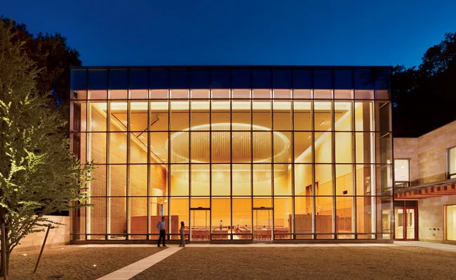Framed by landscaped grounds and the evening sky, the temple&rsquo;s north-facing window wall reveals a soaring sanctuary dramatically illuminated by a luminous mesh circle of light and a floating cei