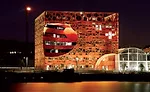 Located in a former dockland, the new commercial building is clad in an aluminum scrim whose pattern was inspired by the movement of water.