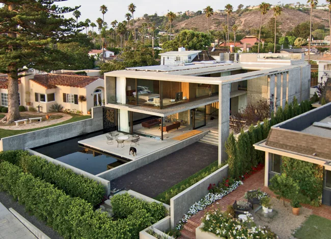 An outdoor room with a lap pool, reflecting pool, and seating areas wraps the house on the ocean-facing side. The hedge-lined walkway softens the transition between the street and the entry and helps